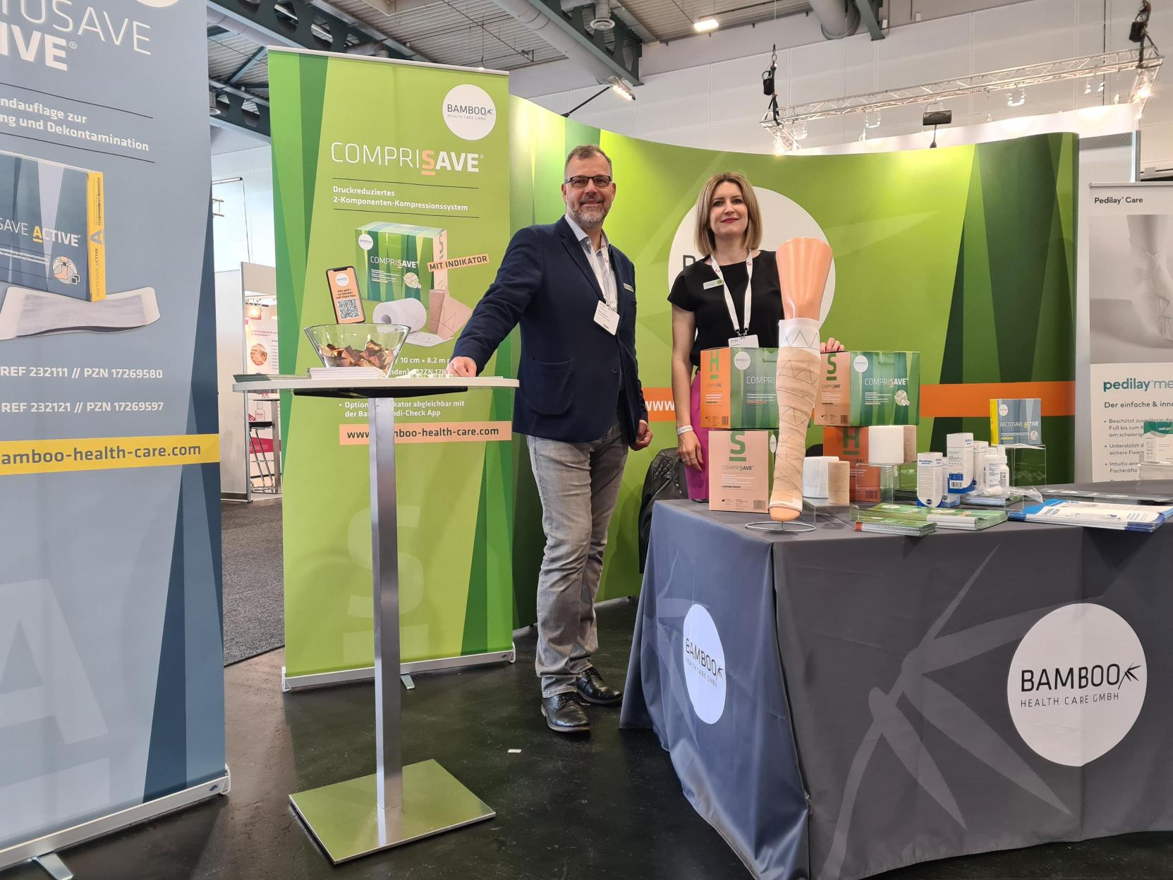 Unsere Außendienst Mitarbeiter Bernd und Tina stehend an unserem Messestand mit einer Auswahl von Bamboo Produktansichtsexemplaren. | © Bamboo Health Care GmbH