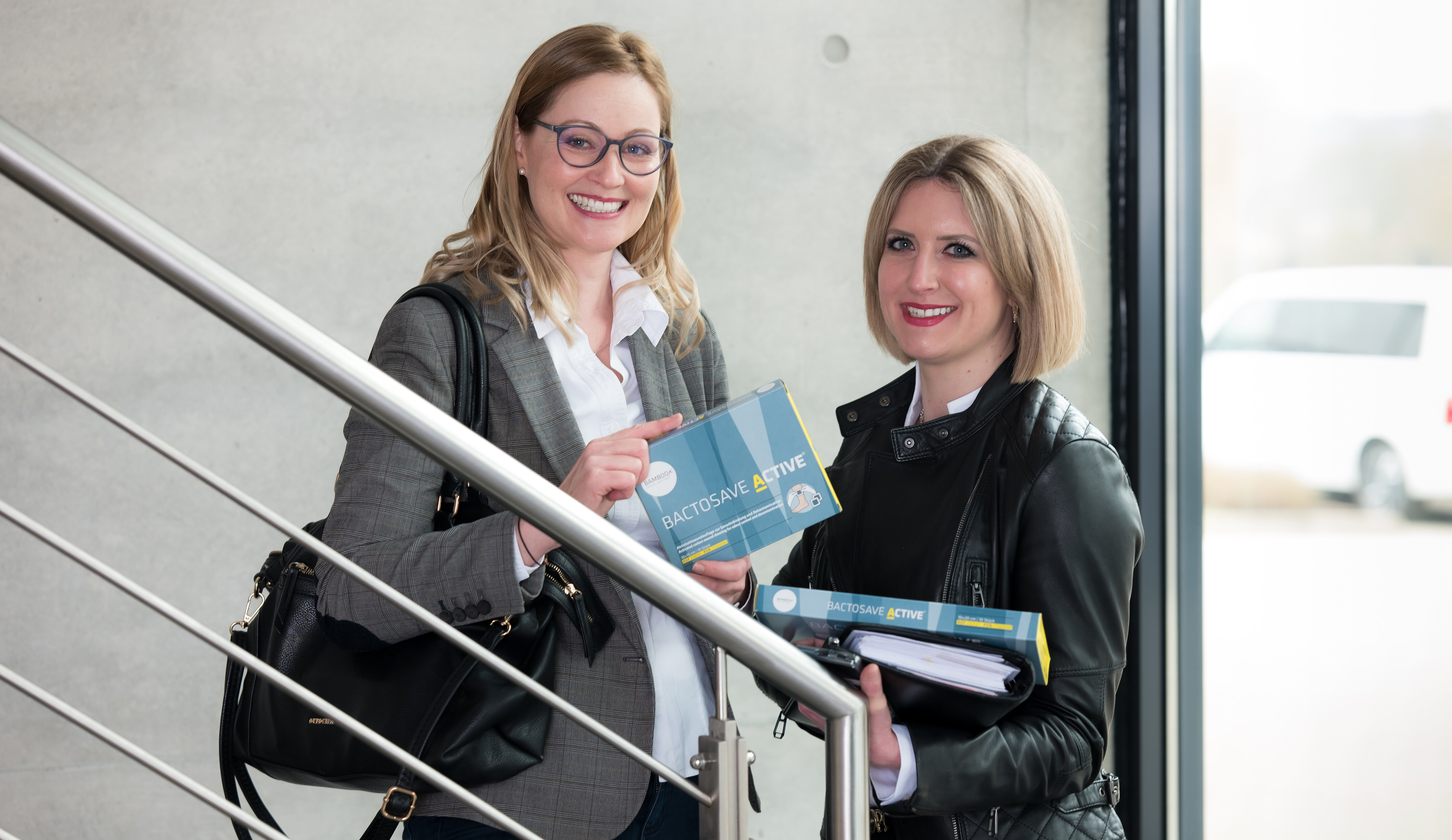 Unsere lächelnden Mitarbeiterinnen Karin und Tina mit BactoSave Active Verpackungen in  der Hand, stehend am Treppenaufgang im Bürogebäude. | © Bamboo Health Care GmbH