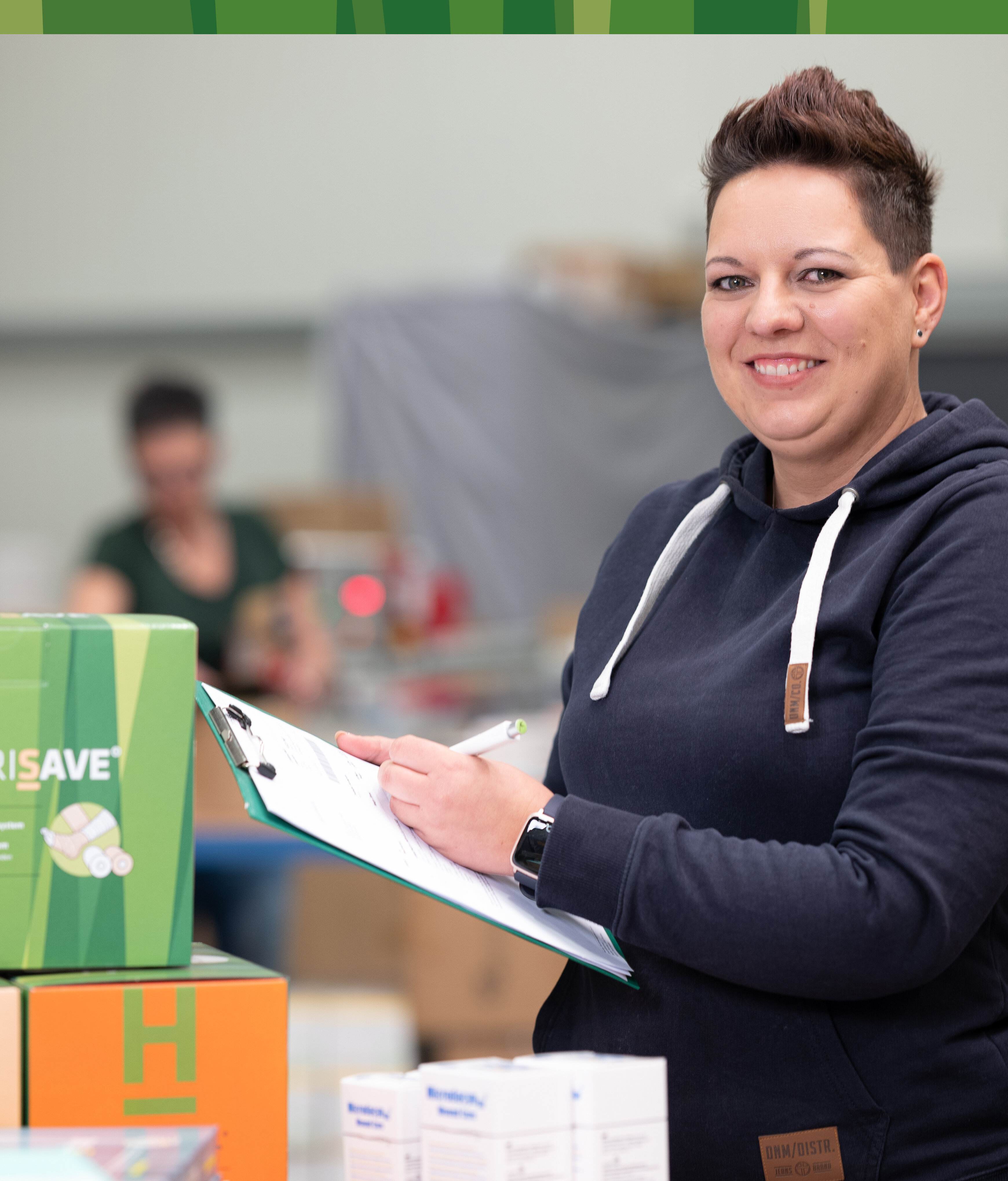 Unsere lächelnde Mitarbeiterin Heike kontrolliert mit einem Klemmbrett und Kugelschreiber in der Hand die vor ihr auf dem Tisch liegende ausgehende Ware im Lager. | © Bamboo Health Care GmbH