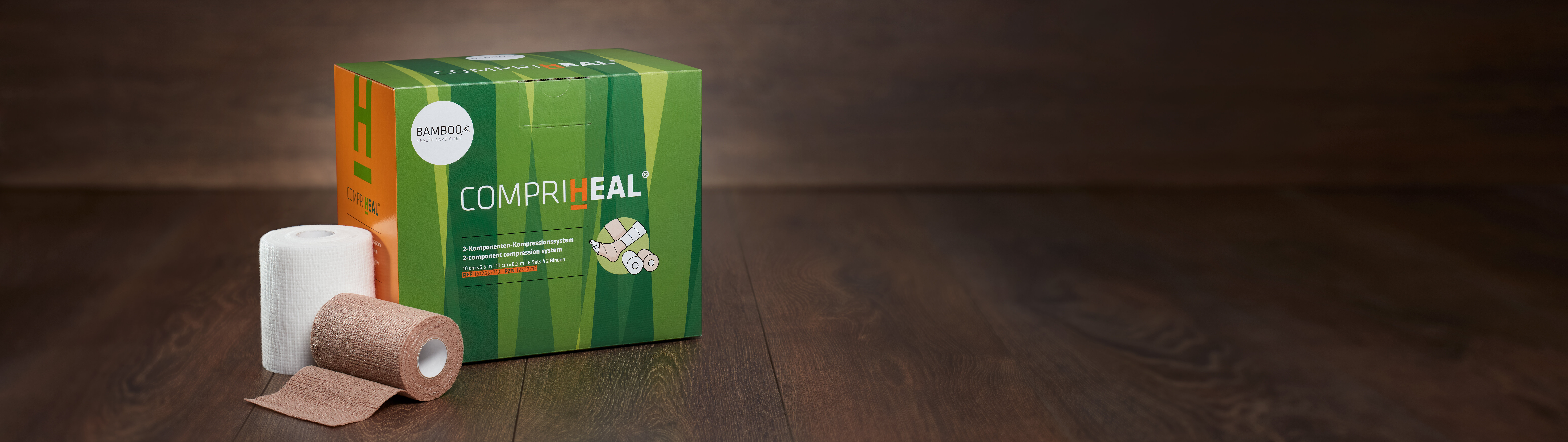 Die Verpackung von Compri-Heal® stehend mit einem Set Kompressionsbinden im Vordergrund auf einem dunklen Holzboden. | © Bamboo Health Care GmbH