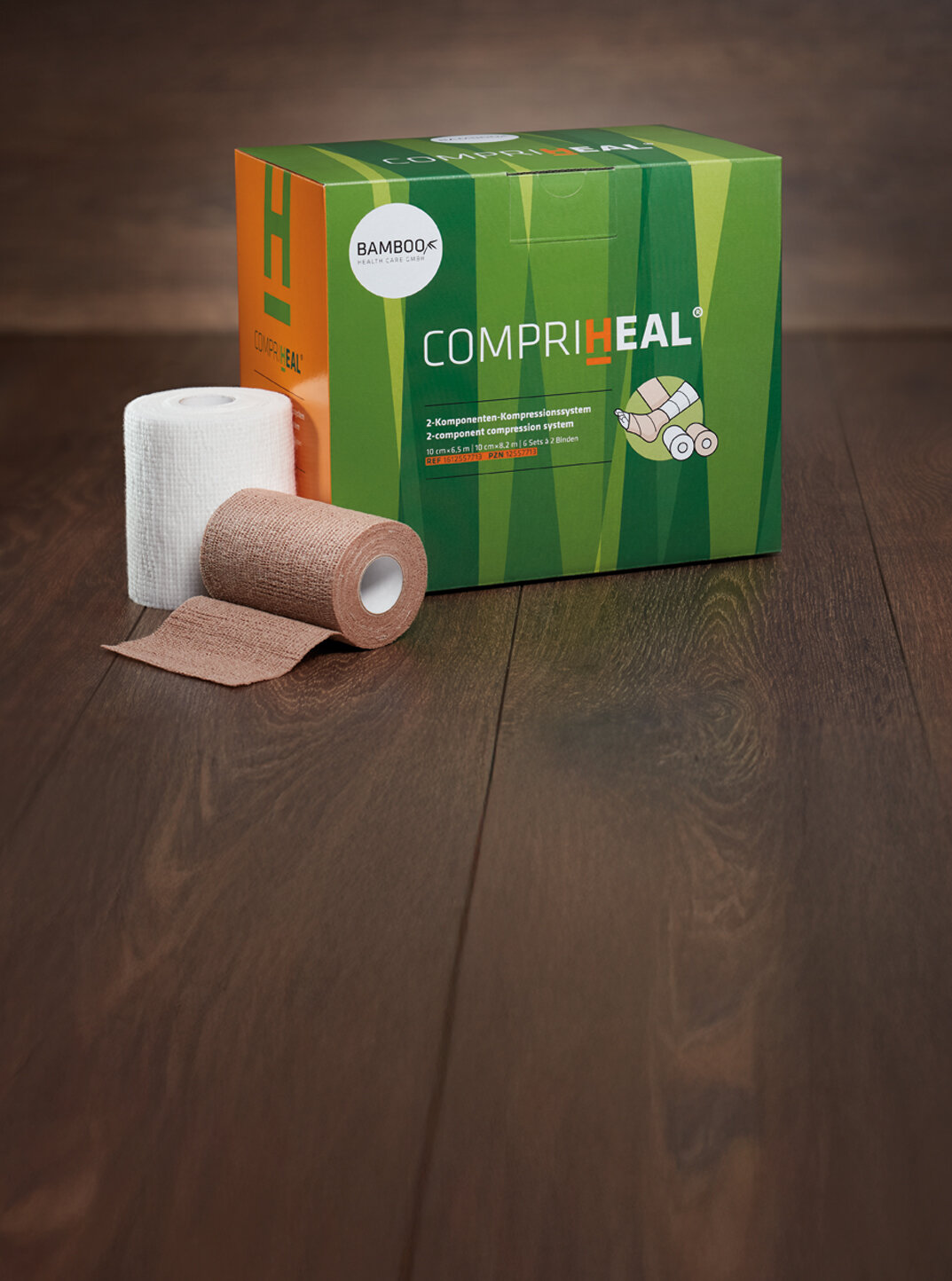 Die Verpackung von Compri-Heal® stehend mit einem Set Kompressionsbinden im Vordergrund auf einem dunklen Holzboden. | © Bamboo Health Care GmbH
