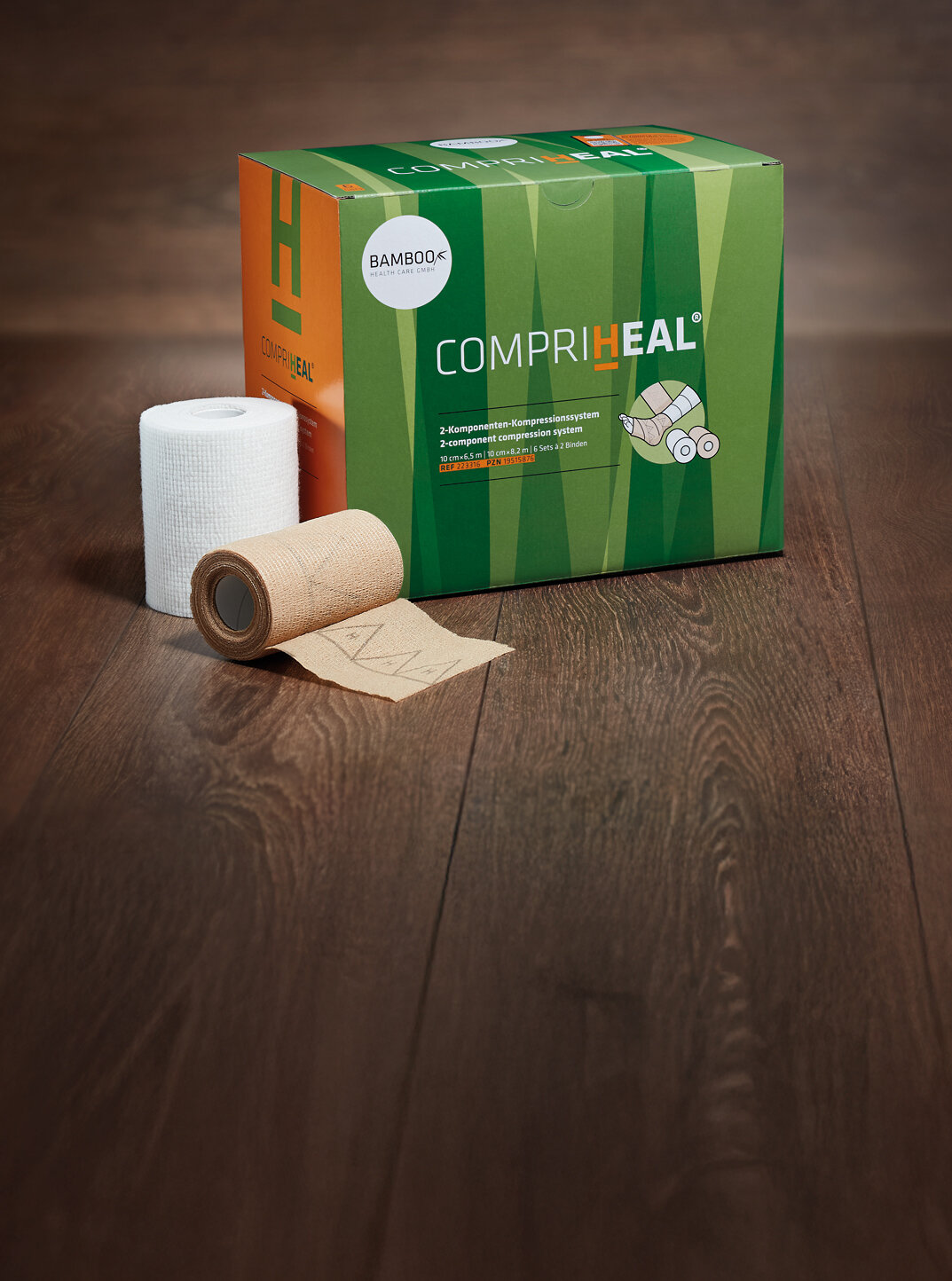 Die Verpackung von CompriHeal® stehend mit einem Set Kompressionsbinden im Vordergrund auf einem dunklen Holzboden. | © Bamboo Health Care GmbH