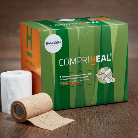 Die Verpackung von CompriHeal® stehend mit einem Set Kompressionsbinden im Vordergrund auf einem dunklen Holzboden. | © Bamboo Health Care GmbH