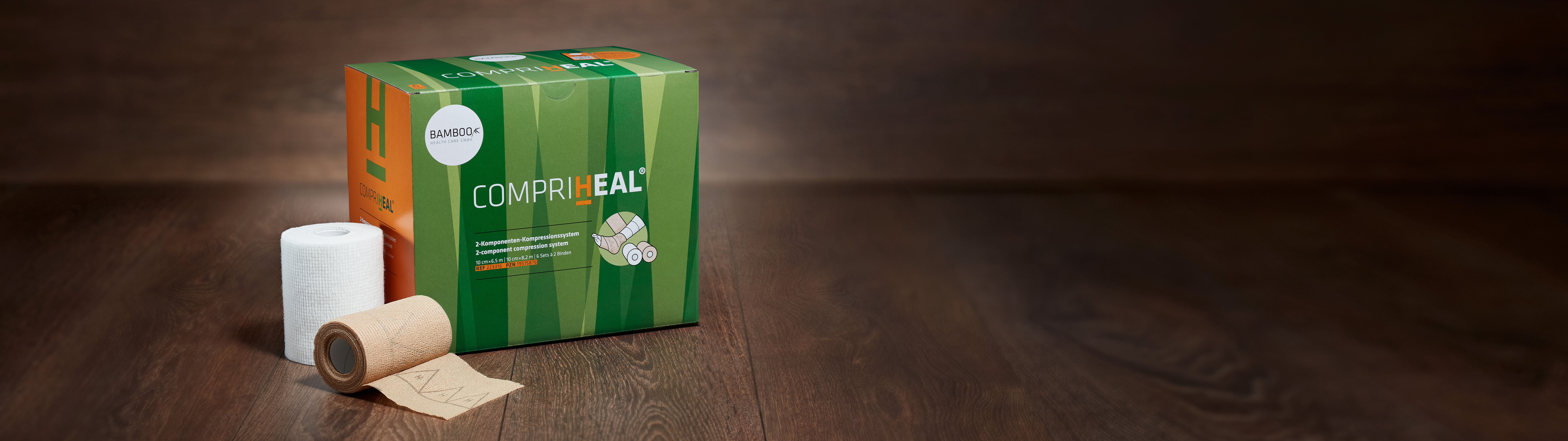 Die Verpackung von CompriHeal® stehend mit einem Set Kompressionsbinden im Vordergrund auf einem dunklen Holzboden. | © Bamboo Health Care GmbH