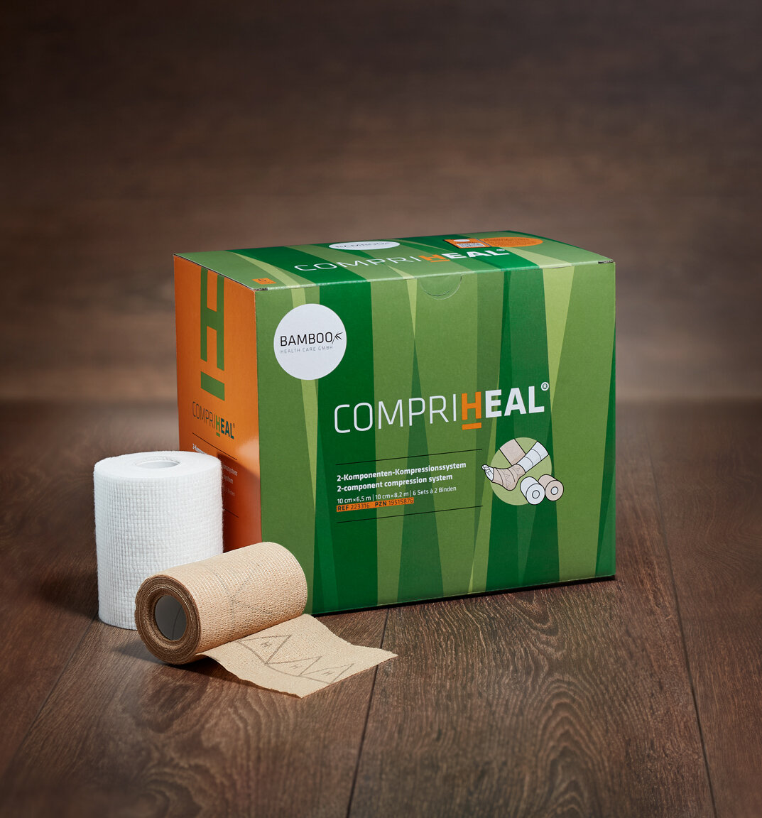 Die Verpackung von CompriHeal® mit der weißen gepolsterten Kurzzug- und der braunen dauerelastischen kohäsiven Langzugbinde im Vordergrund auf dunklem Holzboden. | © Bamboo Health Care GmbH