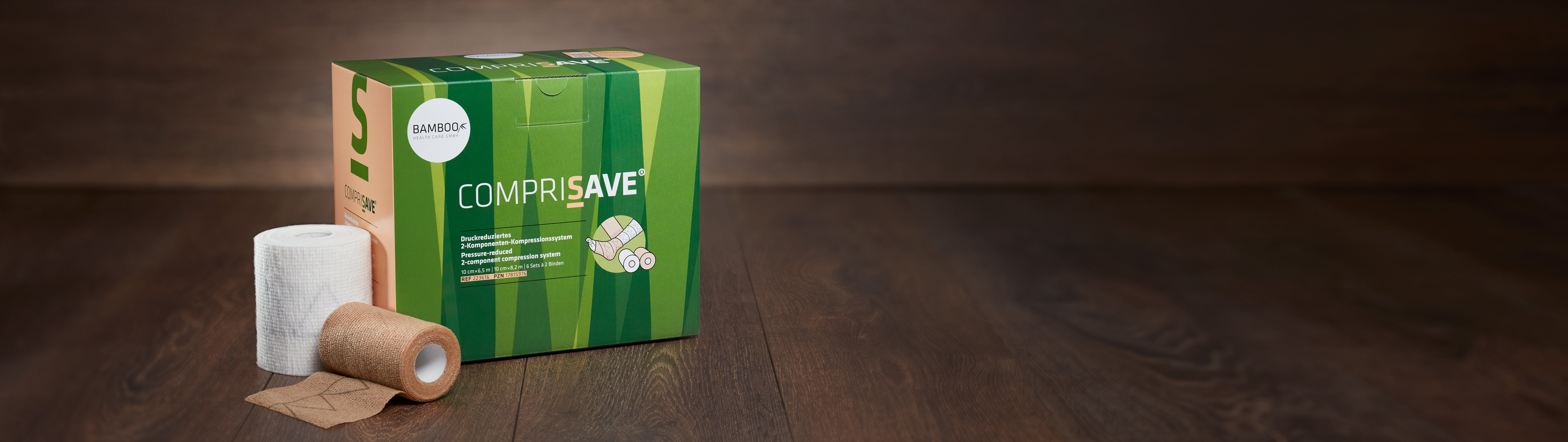 Die Verpackung von CompriSave® stehend mit einem Set Kompressionsbinden im Vordergrund auf einem dunklen Holzboden. | © Bamboo Health Care GmbH