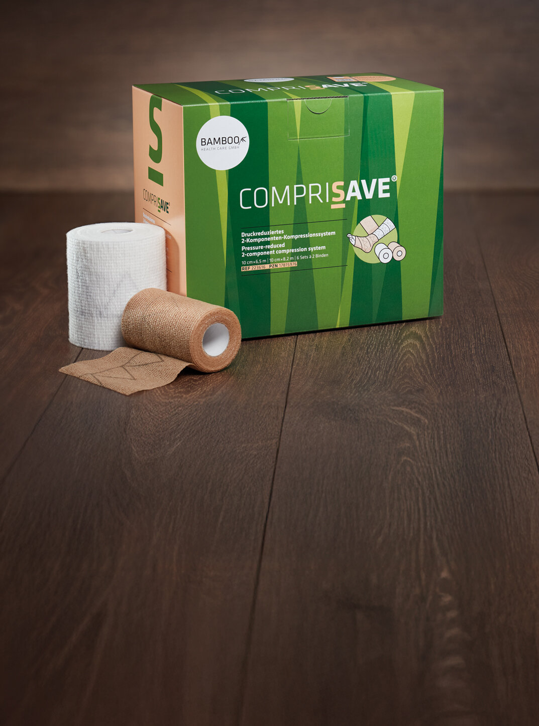 Die Verpackung von CompriSave® stehend mit einem Set Kompressionsbinden im Vordergrund auf einem dunklen Holzboden. | © Bamboo Health Care GmbH