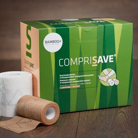 Die Verpackung von CompriSave® stehend mit einem Set Kompressionsbinden im Vordergrund auf einem dunklen Holzboden. | © Bamboo Health Care GmbH