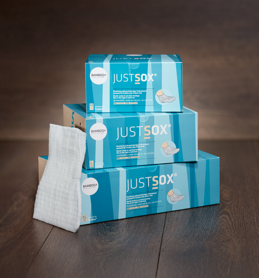 Drei Verpackungsgrößen von JustSox® mit einem JustSox® Fußverband im Vordergrund auf dunklem Holzboden. | © Bamboo Health Care GmbH