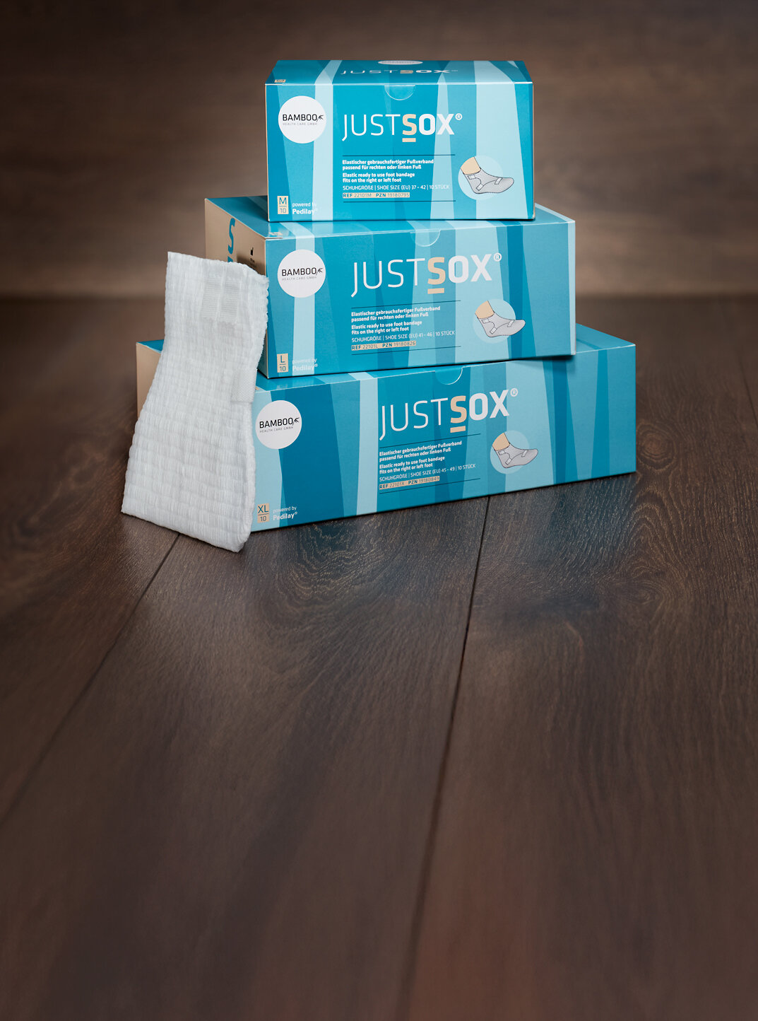 Zwei unterschiedliche Verpackungsgrößen von JustSox® stehend mit zwei Fußverbänden im Vordergrund auf einem dunklen Holzboden. | © Bamboo Health Care GmbH