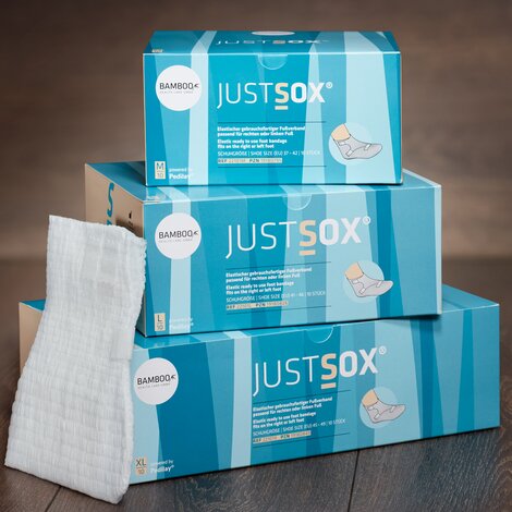 Zwei unterschiedliche Verpackungsgrößen von JustSox® stehend mit zwei Fußverbänden im Vordergrund auf einem dunklen Holzboden. | © Bamboo Health Care GmbH