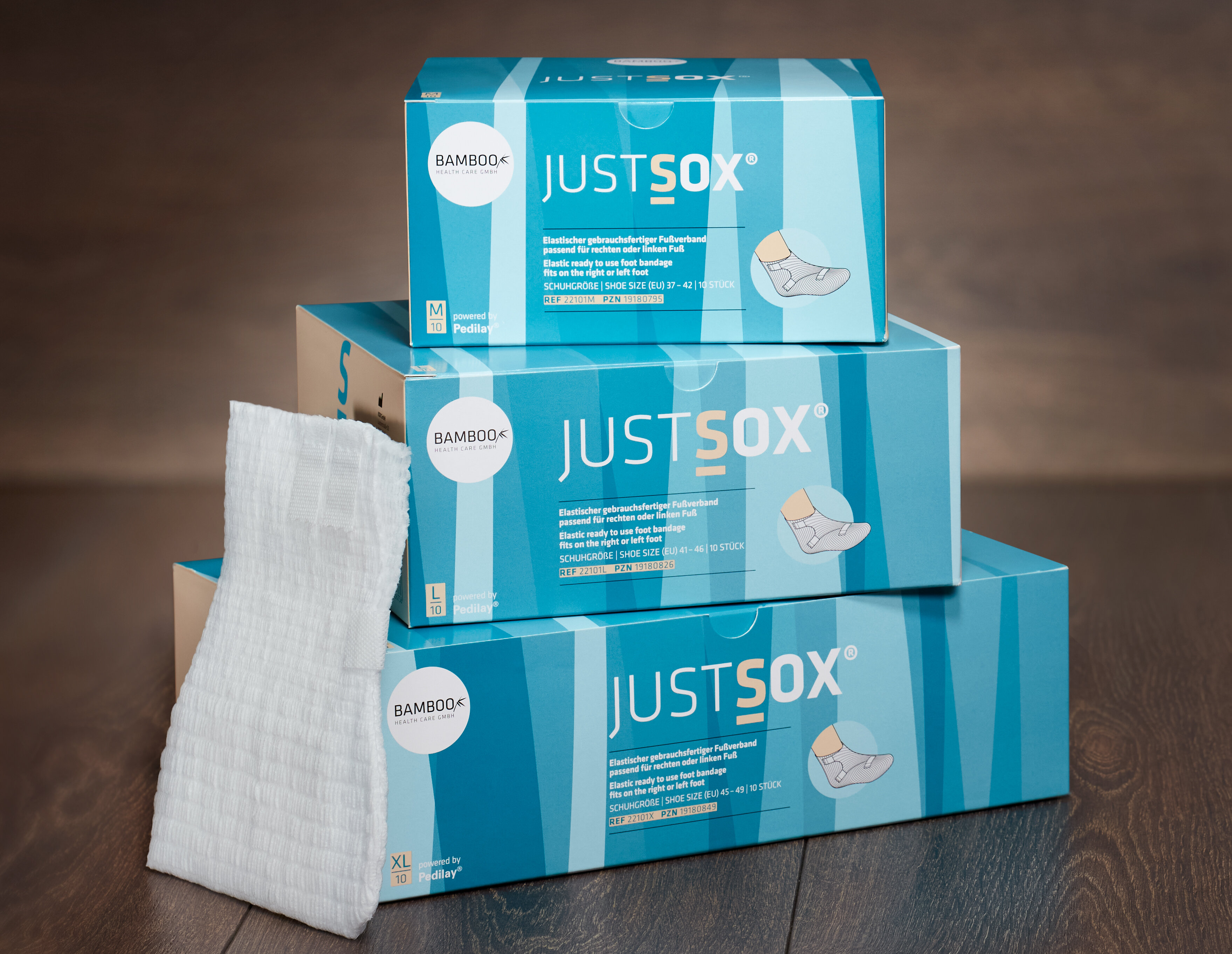 Zwei unterschiedliche Verpackungsgrößen von JustSox® stehend mit zwei Fußverbänden im Vordergrund auf einem dunklen Holzboden. | © Bamboo Health Care GmbH