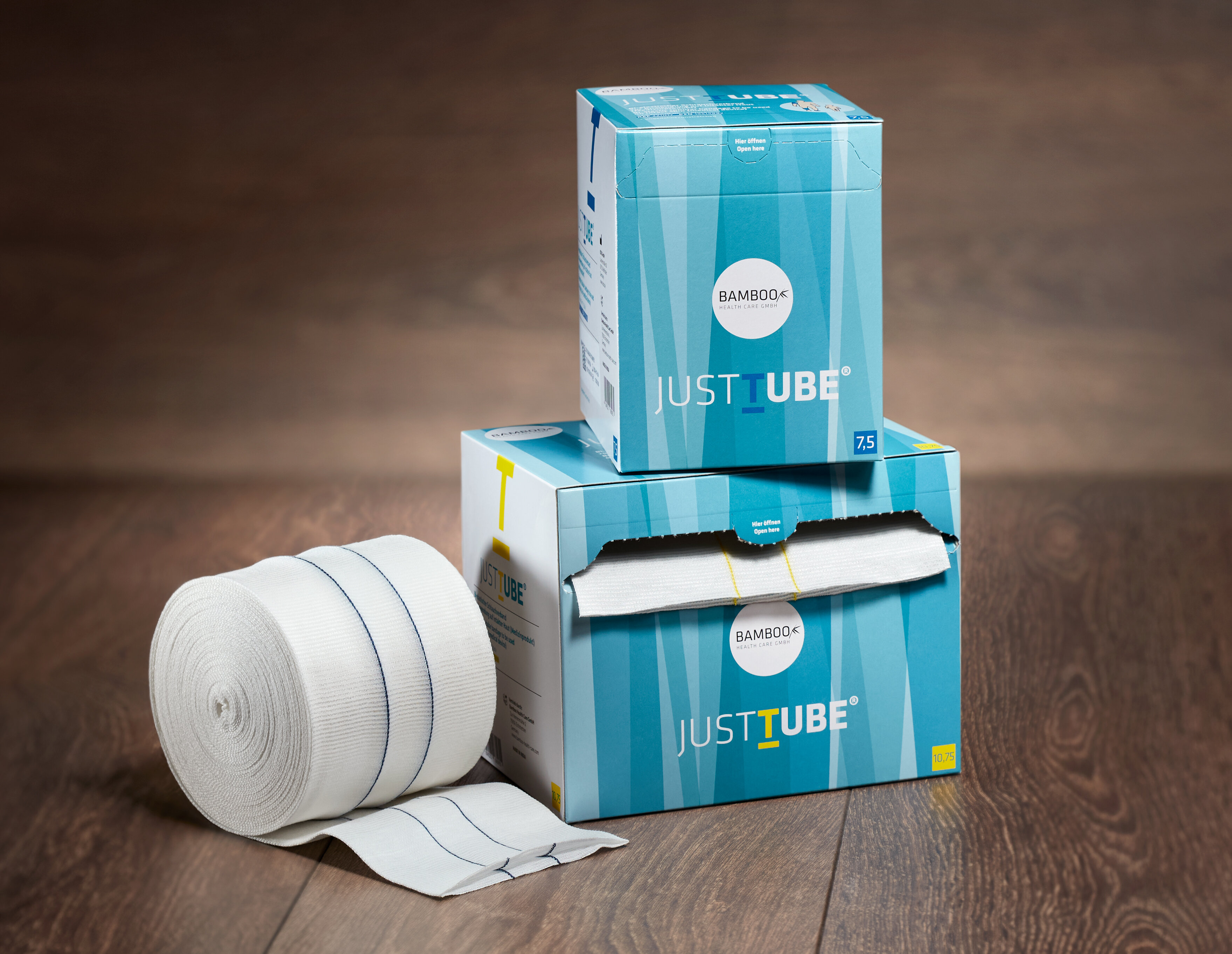 Zwei unterschiedliche Verpackungsgrößen von JustTube® aufeinander gestapelt mit Schlauchverband links im Vordergrund auf einem dunklen Holzboden. | © Bamboo Health Care GmbH