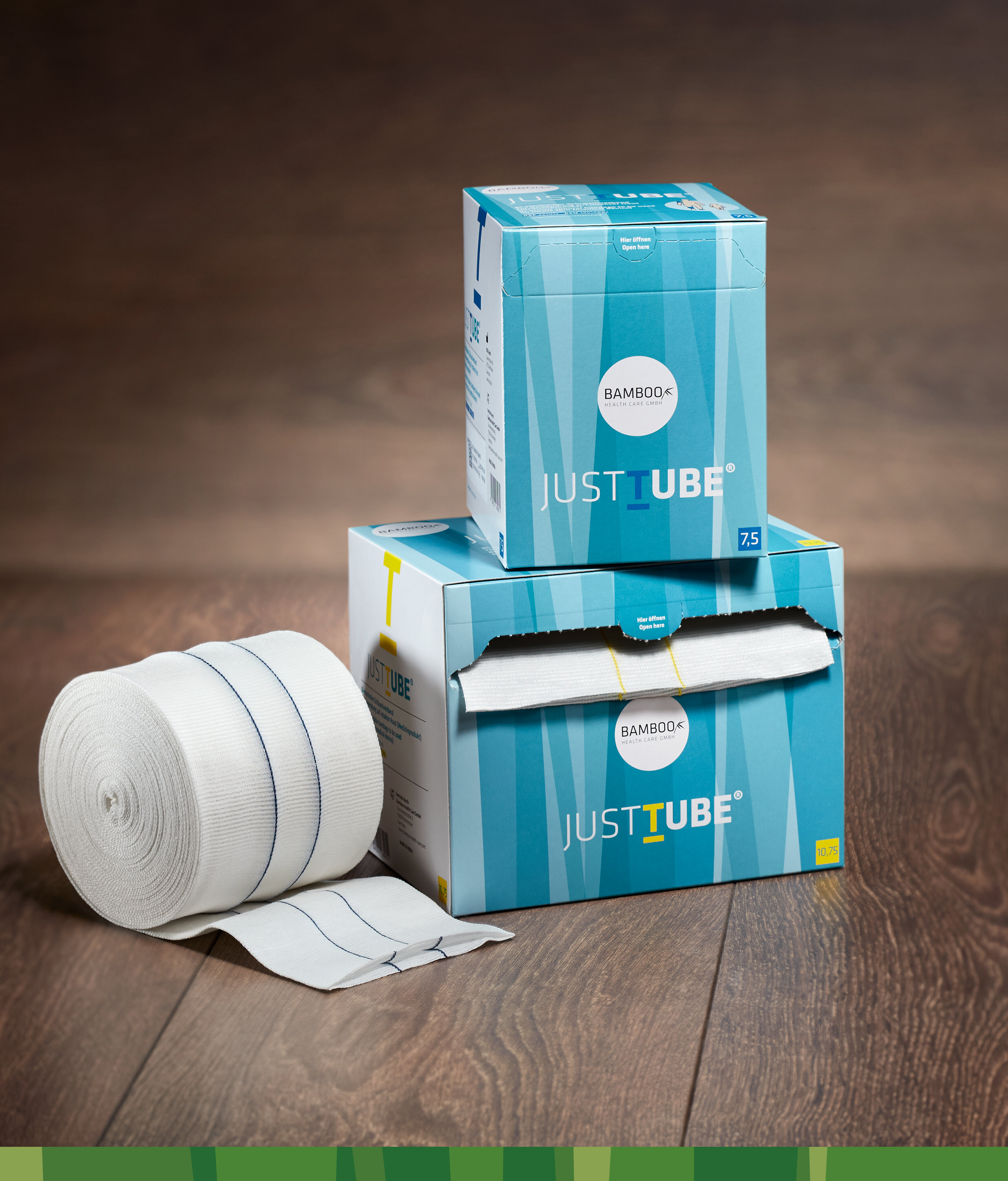 Zwei unterschiedliche Verpackungsgrößen von JustTube® aufeinander gestapelt mit Schlauchverband links im Vordergrund auf einem dunklen Holzboden. | © Bamboo Health Care GmbH