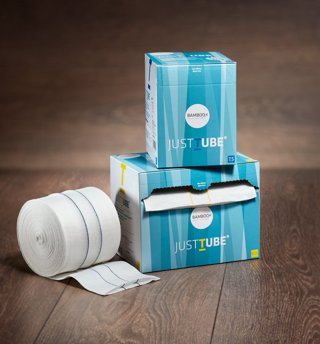 Die zwei Verpackungsgrößen von JustTube® mit dem JustTube® Schlauchverband in der Größe >7,5 cm×10 m im Vordergrund auf dunklem Holzboden. | © Bamboo Health Care GmbH