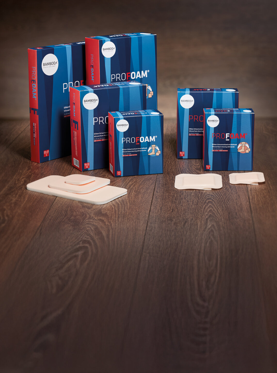 Alle sechs Verpackungsgrößen von ProFoam® mit und ohne Haftrand nebeneinander gestellt mit passendem Schaumverband im Vordergrund auf einem dunklen Holzboden. | © Bamboo Health Care GmbH