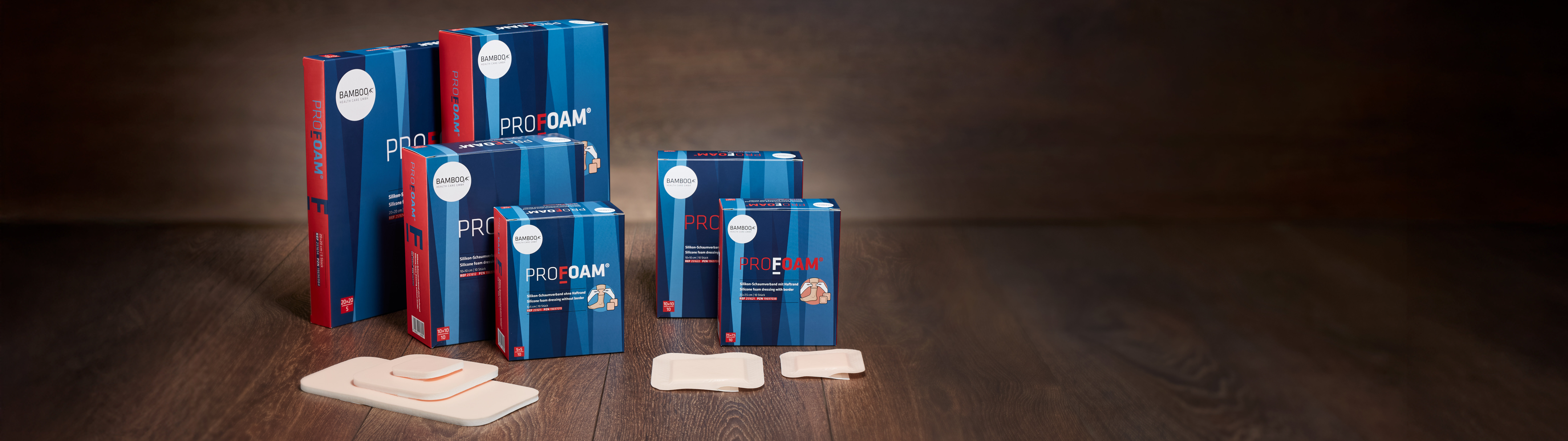 Alle sechs Verpackungsgrößen von ProFoam® mit und ohne Haftrand nebeneinander gestellt mit passendem Schaumverband im Vordergrund auf einem dunklen Holzboden. | © Bamboo Health Care GmbH
