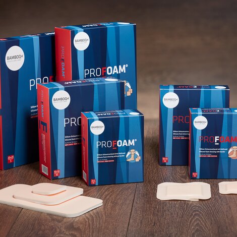 Alle sechs Verpackungsgrößen von ProFoam® mit und ohne Haftrand nebeneinander gestellt mit passendem Schaumverband im Vordergrund auf einem dunklen Holzboden. | © Bamboo Health Care GmbH