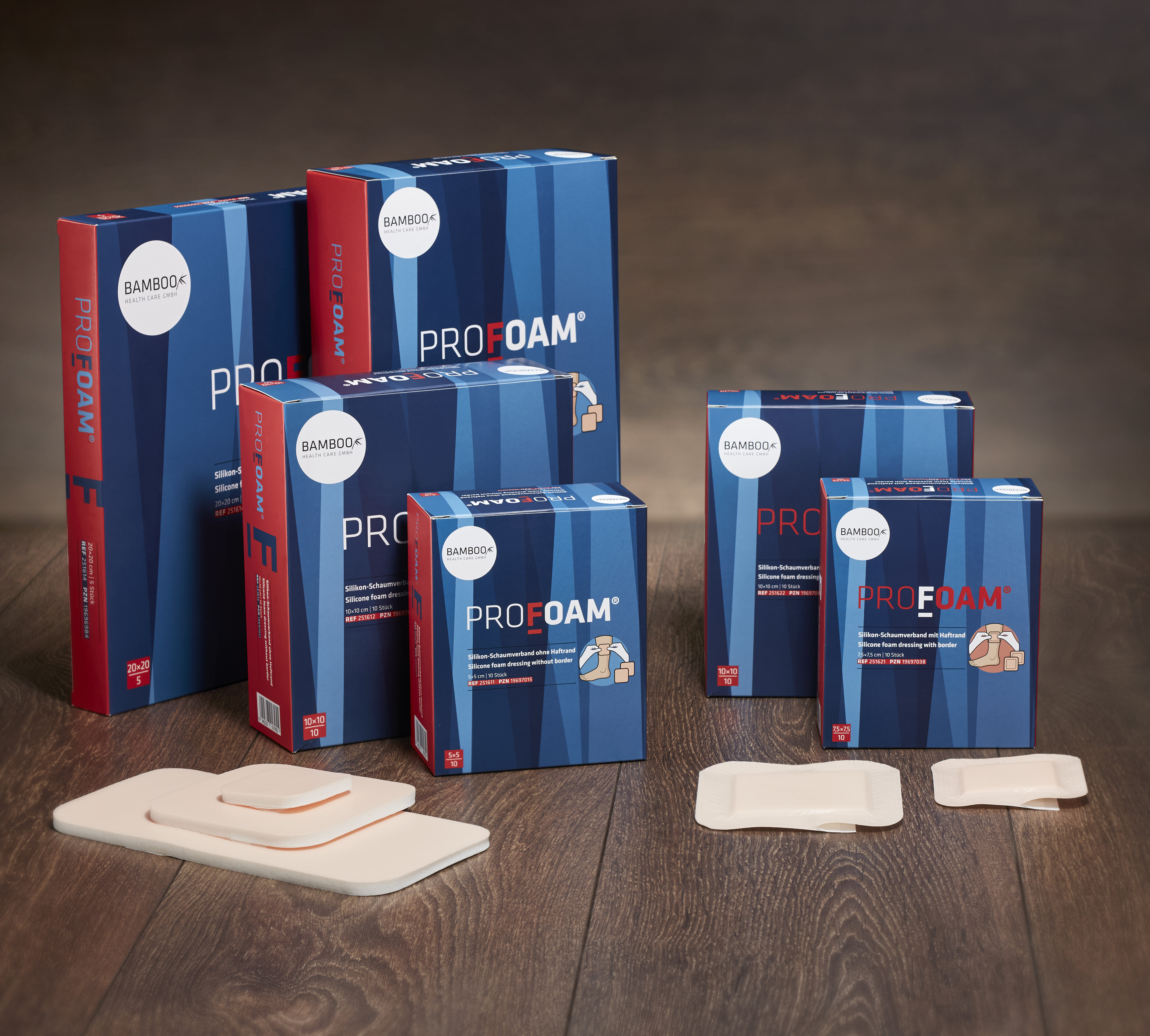 Alle sechs Verpackungsgrößen von ProFoam® mit und ohne Haftrand nebeneinander gestellt mit passendem Schaumverband im Vordergrund auf einem dunklen Holzboden. | © Bamboo Health Care GmbH