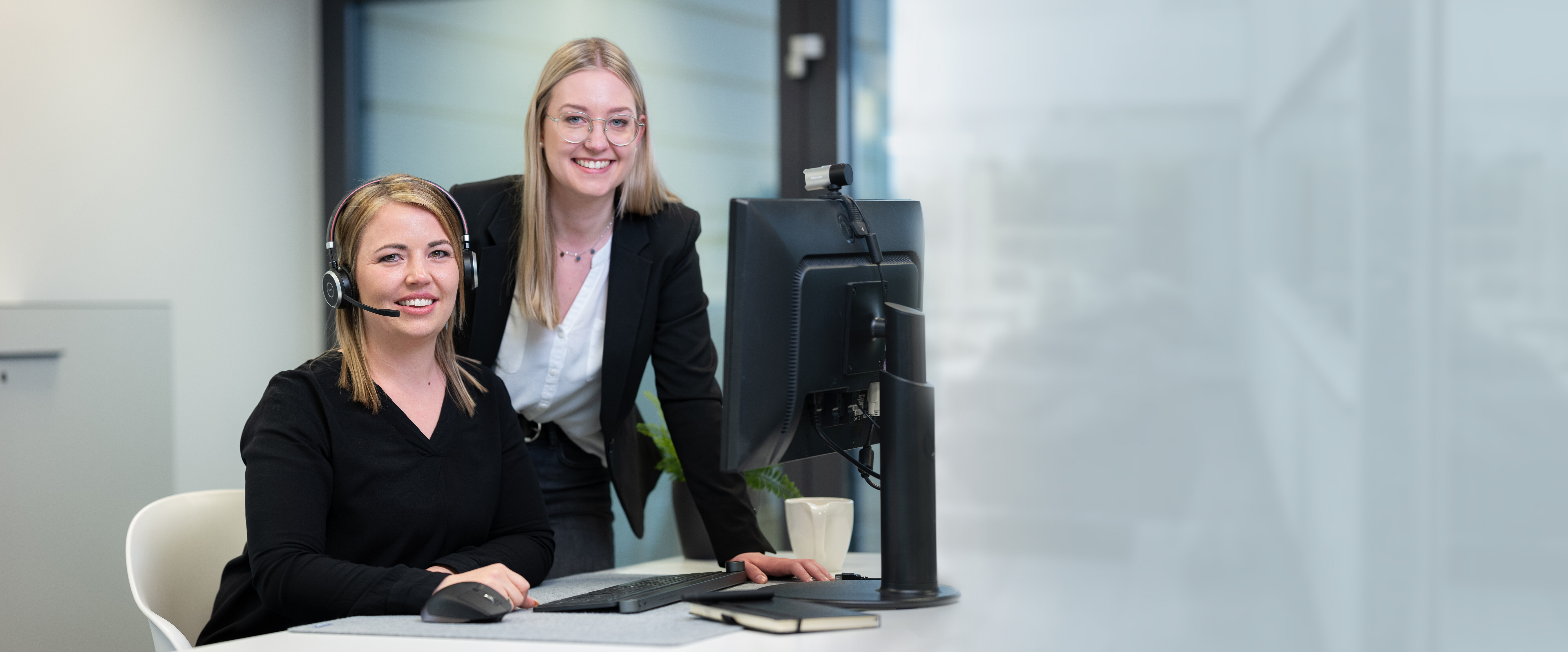 Unsere lächelnden Mitarbeiterinnen Laura mit Headset und Larissa daneben stehend in einer alltäglichen Arbeitssituation gemeinsam vor dem Computerbildschirm. | © Bamboo Health Care GmbH