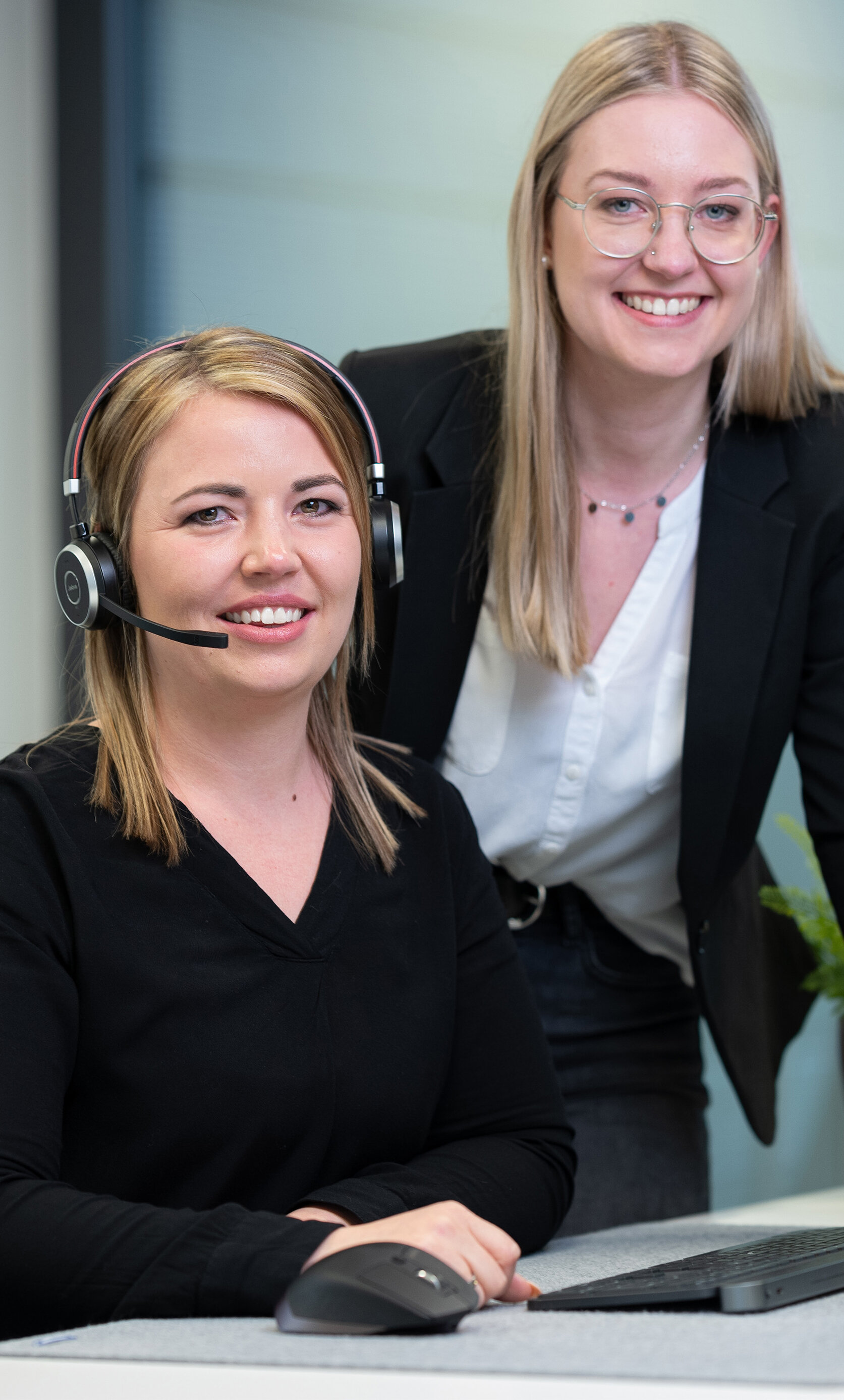 Unsere lächelnden Mitarbeiterinnen Laura mit Headset und Larissa daneben stehend in einer alltäglichen Arbeitssituation gemeinsam vor dem Computerbildschirm. | © Bamboo Health Care GmbH
