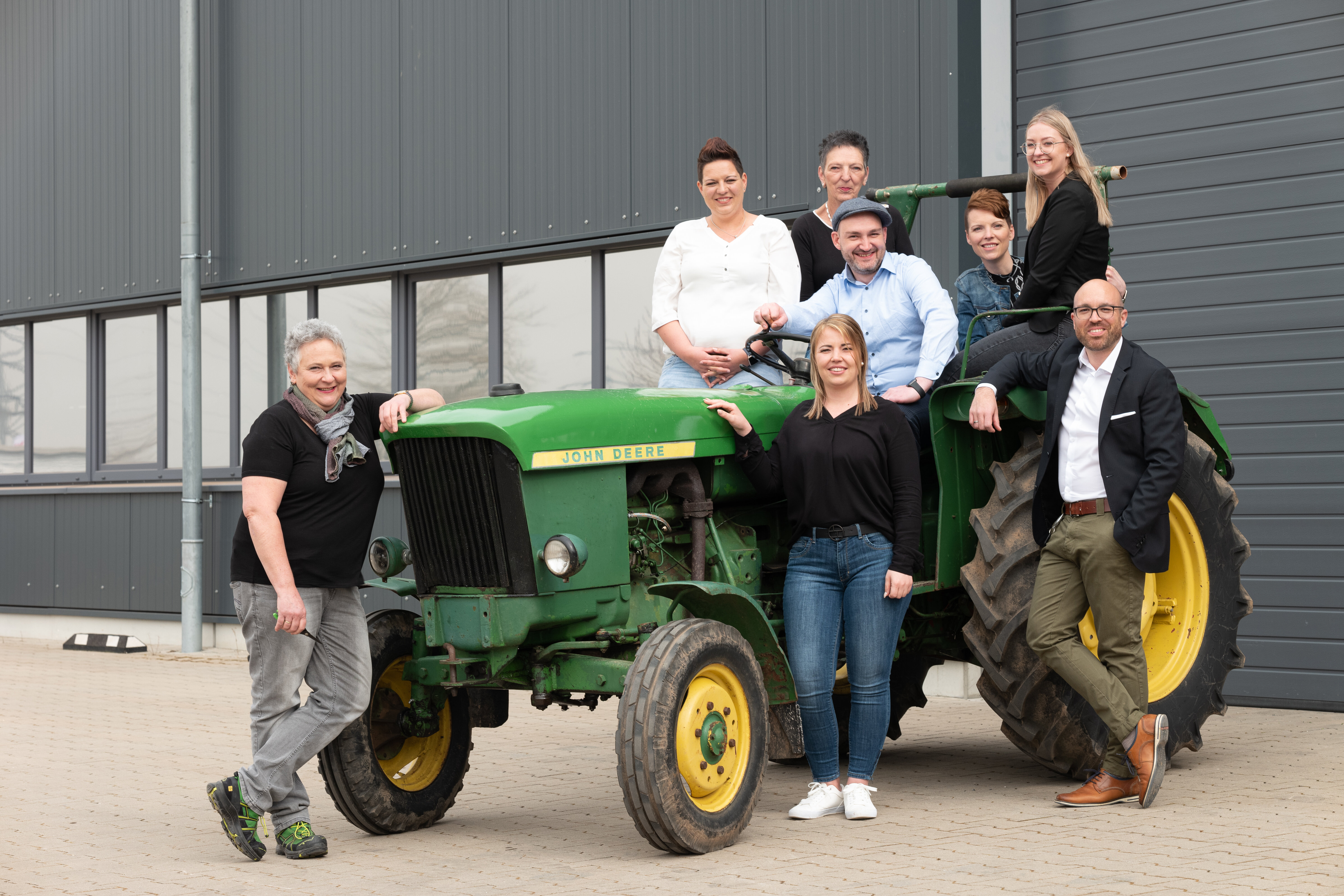 Unser lächelndes Innendienst- und Lager-Team von links nach rechts: Elisabeth, Heike, Laura, Michaela, Kolja, Brigitte, Larissa und Felix, sitzend und stehend auf bzw. um einen grünen Traktor vor der geschlossenen Lagerhalle.